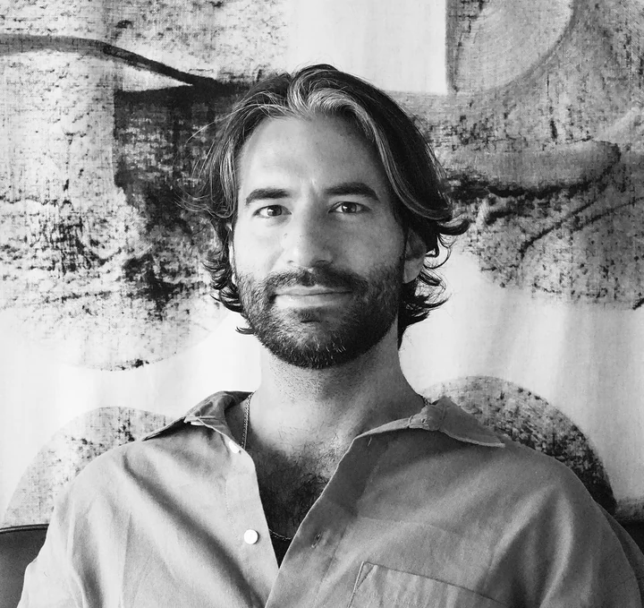Black-and-white portrait of Malek Najm Ghaleb, smiling, in front of a large ink artwork.